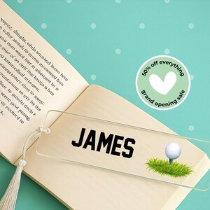 May include: Clear acrylic bookmark with the name "JAMES" in bold black letters. A golf ball on a tee is depicted on a patch of green grass. The bookmark is placed on an open book with a tassel.