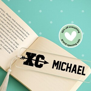 May include: Clear acrylic bookmark with the text "XC MICHAEL" in black. The bookmark has a tassel and is placed on an open book. A sticker with a heart and the text "50% off everything grand opening sale" is also visible.