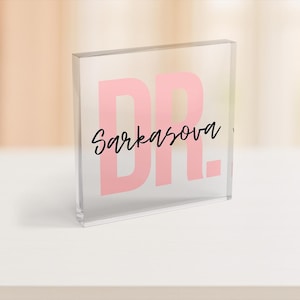 May include: A clear acrylic block with the word "DR." in pink, and the name "Sarkasova" in black script. The block is on a white surface, with a blurred background.
