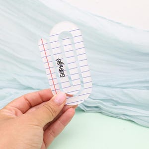 May include: A clear acrylic paperclip with a notebook paper design. The paperclip has blue lines and a red margin, with the name "George" printed in black script. The paperclip is held against a light blue background.
