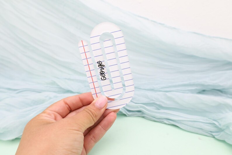 May include: A clear acrylic paperclip with a notebook paper design. The paperclip has blue lines and a red margin, with the name "George" printed in black script. The paperclip is held against a light blue background.