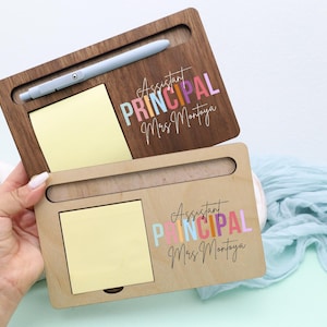 May include: Two wooden desk organizers, one dark brown and one light brown, each with a slot for a pen, a yellow sticky note pad, and the text "Assistant Principal Mrs. Montoya" in colorful letters. A light blue pen rests in the top organizer.