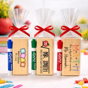 May include: Three gift sets for teachers, each with a wooden block and a marker. The blocks have math-themed designs and teacher names. The sets are wrapped in clear cellophane and tied with red ribbons. The markers are blue, green, and red.