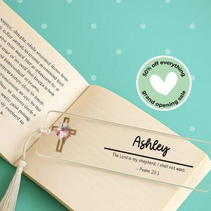 May include: Clear acrylic bookmark with a gold cross adorned with flowers. The name "Ashley" is written in black script, along with the Bible verse Psalm 23:1. A tassel hangs from the top. A "50% off everything" sale sticker is in the background.