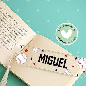 May include: Clear acrylic bookmark with the name "MIGUEL" in black block letters. Baseball graphics, including red stars, baseballs, and blue lightning bolts, decorate the bookmark. A tassel hangs from the top.