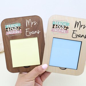 May include: Two wooden memo holders, one dark brown and one light, each with a rectangular opening for notes. The text "TEACH LOVE INSPIRE" is above the opening, and "Mrs. Evans" is written below. One has a yellow note, the other a blue one.