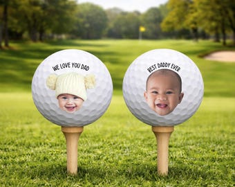 Può includere: Due palline da golf su tee di legno, ognuna con la faccia di un bambino. Una pallina dice "WE LOVE YOU DAD" e il bambino indossa un cappello. L'altra dice "BEST DADDY EVER". Le palline da golf sono su un campo da golf verde.