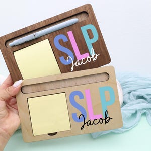 May include: Two wooden desk organizers, one dark brown and one light brown, each with a slot for a pen, a yellow sticky note pad, and the name "Jacob" with "SLP" in colorful letters. A light blue pen rests in the top organizer.
