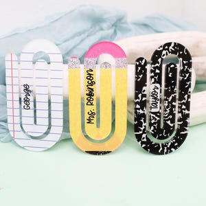 May include: Three decorative paperclip-shaped bookmarks. One is white with blue lines and the name "George". The second is yellow with a pink top and the name "Mrs. Robinson". The third is black and white with the name "Taylor".