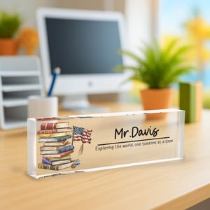 A clear glass block on a wooden desk displays a stack of books with an American flag and the name "Mr. Davis" written on it.