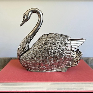 May include: A silver-toned metal swan figurine featuring detailed, textured feather patterns across its body and a long, curved neck. The swan is depicted in a resting position with its wings folded, showcasing a metallic finish and intricate craftsmanship.