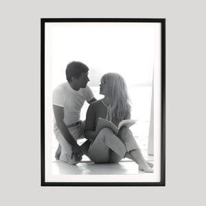 May include: Black and white framed photograph of a man and woman. The man is kneeling, looking at the woman, who is seated and holding a book. The photograph is in a black frame.