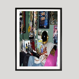 Pode incluir: Impressão emoldurada com duas figuras com capacetes Daft Punk lendo quadrinhos. Uma figura usa um capacete prateado e uma camisa vermelha, a outra um capacete preto e dourado e uma camisa de beisebol. O fundo inclui cartazes de filmes.