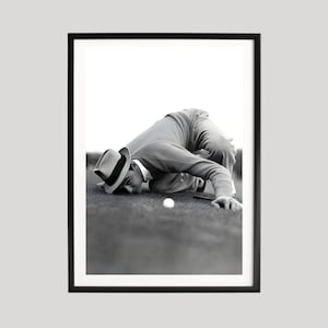 May include: Framed monochrome print of a person on a golf course, focused on a golf ball. The person is wearing a hat, sweater, and trousers. The image is a medium shot, with a black frame.