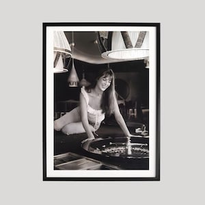 Op de afbeelding: Zwart-wit ingelijste foto van een lachende vrouw, leunend op een roulettetafel. De afbeelding is geplaatst in een zwak verlichte kamer met hanglampen. De vrouw draagt een lichtgekleurde top.
