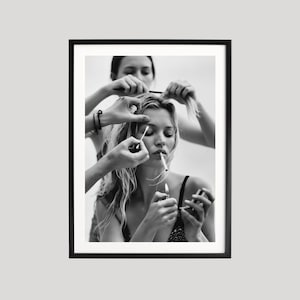 May include: Black and white framed print of a woman with long hair, smoking a cigarette while having her hair and makeup done. Several hands are visible, holding a lighter, comb, and makeup tools. The image has a vintage, editorial feel.
