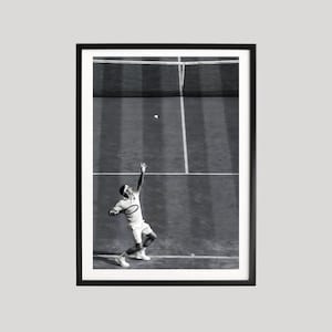 May include: Black and white framed print of a tennis player serving on a grass court. The player is wearing white athletic attire and is mid-serve, with the ball in the air. The net and court lines are visible.