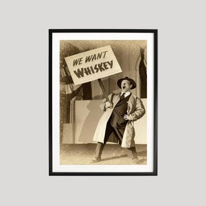 May include: A framed sepia-toned print featuring a man in a coat and hat holding a sign that reads "WE WANT WHISKEY." The man is shouting with his mouth open. The print is in a black frame.