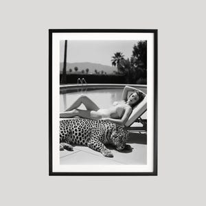 May include: Black and white framed print featuring a woman reclining on a lounge chair next to a leopard. The woman is wearing a swimsuit. The background includes a pool, palm trees, and mountains.