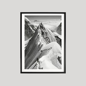 May include: Black and white framed photograph of a mountain range. The image captures a snow-covered mountain ridge with a person climbing along the edge. The sky is filled with clouds, and the photograph is framed in a black border.