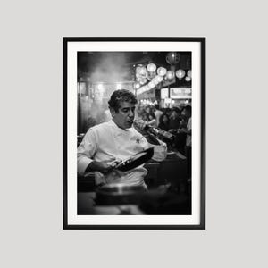 Peut inclure: Tirage encadr&eacute; en noir et blanc d'un chef cuisinier en veste blanche, tenant une po&ecirc;le et une bouteille. Le chef est dans un restaurant anim&eacute;, avec d'autres personnes en arri&egrave;re-plan. L'image est un portrait.