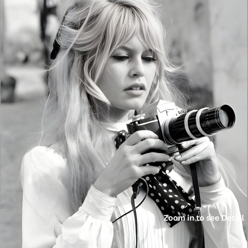 May include: Monochrome image of a person holding a vintage camera with a long lens. The camera features white rings and a black strap with white polka dots. The person is wearing a white blouse and has long blonde hair.