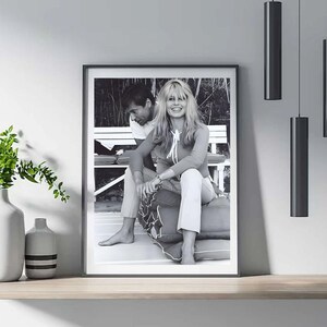 May include: Framed black and white photograph featuring a seated couple. The woman is smiling, wearing a long-sleeved top and pants. The man is seated next to her. The photograph is displayed on a wooden shelf.