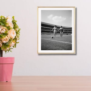 K&ouml;nnte beinhalten: Gerahmtes Schwarz-Wei&szlig;-Foto von zwei Personen, die auf einem Fu&szlig;ballfeld laufen. Das Foto ist aus einer niedrigen Perspektive aufgenommen, mit dem Stadion im Hintergrund. Ein Fu&szlig;ball liegt auf dem Feld.