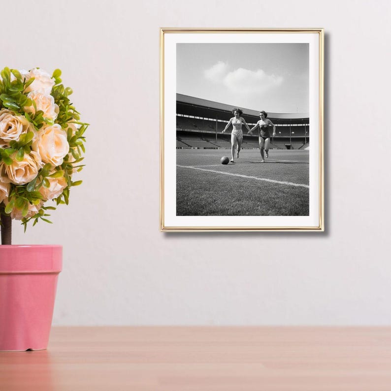 K&ouml;nnte beinhalten: Gerahmtes Schwarz-Wei&szlig;-Foto von zwei Personen, die auf einem Fu&szlig;ballfeld laufen. Das Foto ist aus einer niedrigen Perspektive aufgenommen, mit dem Stadion im Hintergrund. Ein Fu&szlig;ball liegt auf dem Feld.
