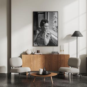 Pu&ograve; includere: Una stampa incorniciata in bianco e nero di Frida Kahlo che fuma, esposta sopra un mobile in legno. La stanza presenta due sedie grigie, un tavolino rotondo e una lampada da terra nera. Include vasi, candele e libri.