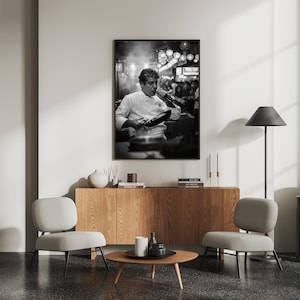 Peut inclure: Impression en noir et blanc d'un chef en veste blanche, go&ucirc;tant un plat. L'arri&egrave;re-plan montre une sc&egrave;ne de rue anim&eacute;e avec des lumi&egrave;res. L'impression est affich&eacute;e au-dessus d'un meuble en bois avec deux chaises grises et une table basse ronde.