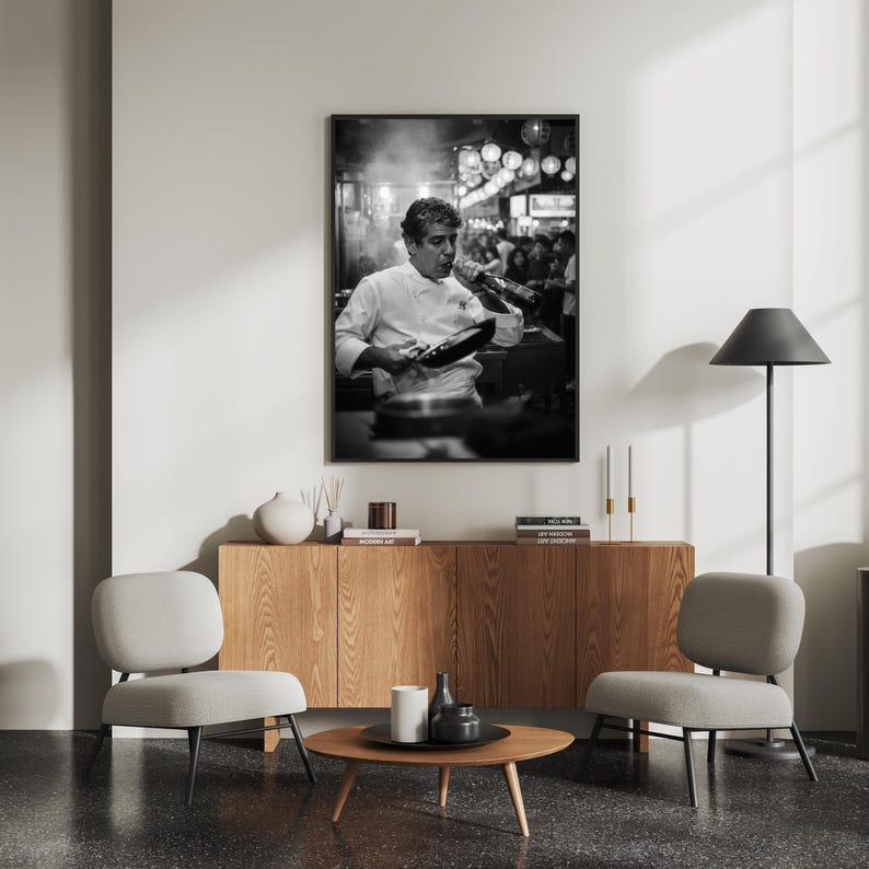 Peut inclure: Impression en noir et blanc d'un chef en veste blanche, go&ucirc;tant un plat. L'arri&egrave;re-plan montre une sc&egrave;ne de rue anim&eacute;e avec des lumi&egrave;res. L'impression est affich&eacute;e au-dessus d'un meuble en bois avec deux chaises grises et une table basse ronde.