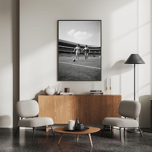K&ouml;nnte beinhalten: Schwarz-wei&szlig; gerahmtes Foto von zwei Personen, die auf einem Fu&szlig;ballfeld laufen. Das Foto h&auml;ngt &uuml;ber einem Holzschrank mit zwei grauen St&uuml;hlen und einem runden Couchtisch. Rechts steht eine schwarze Stehlampe.