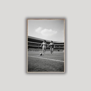 K&ouml;nnte beinhalten: Schwarz-Wei&szlig;-Fotografie von zwei Personen, die auf einem Rasenfeld rennen und sich an den H&auml;nden halten. Ein Fu&szlig;ball liegt auf dem Boden. Der Hintergrund zeigt ein Stadion mit Sitzreihen unter einem bew&ouml;lkten Himmel.