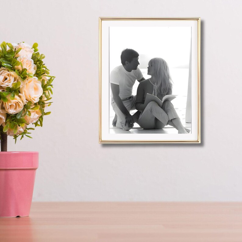 May include: A gold-framed black and white photograph of a couple looking at each other. The photograph is mounted on a white wall. A pink flower pot with a plant is in the foreground. The photograph is a classic piece of art.