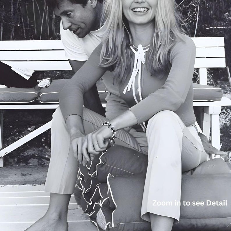 May include: Black and white photograph of a couple sitting on a bench. The woman is wearing a red long-sleeved top with white trim and light-colored pants. She has a bracelet on her wrist and is smiling. The man is wearing a white t-shirt.