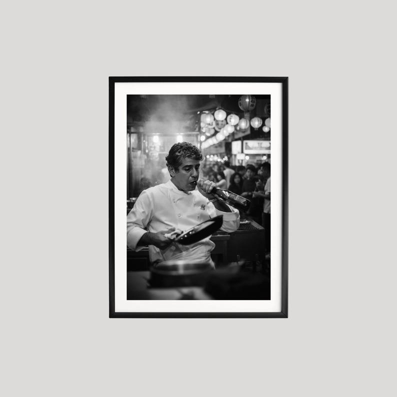 Peut inclure: Tirage encadr&eacute; en noir et blanc d'un chef cuisinier en veste blanche, tenant une po&ecirc;le et une bouteille. Le chef est dans un restaurant anim&eacute;, avec d'autres personnes en arri&egrave;re-plan. L'image est un portrait.