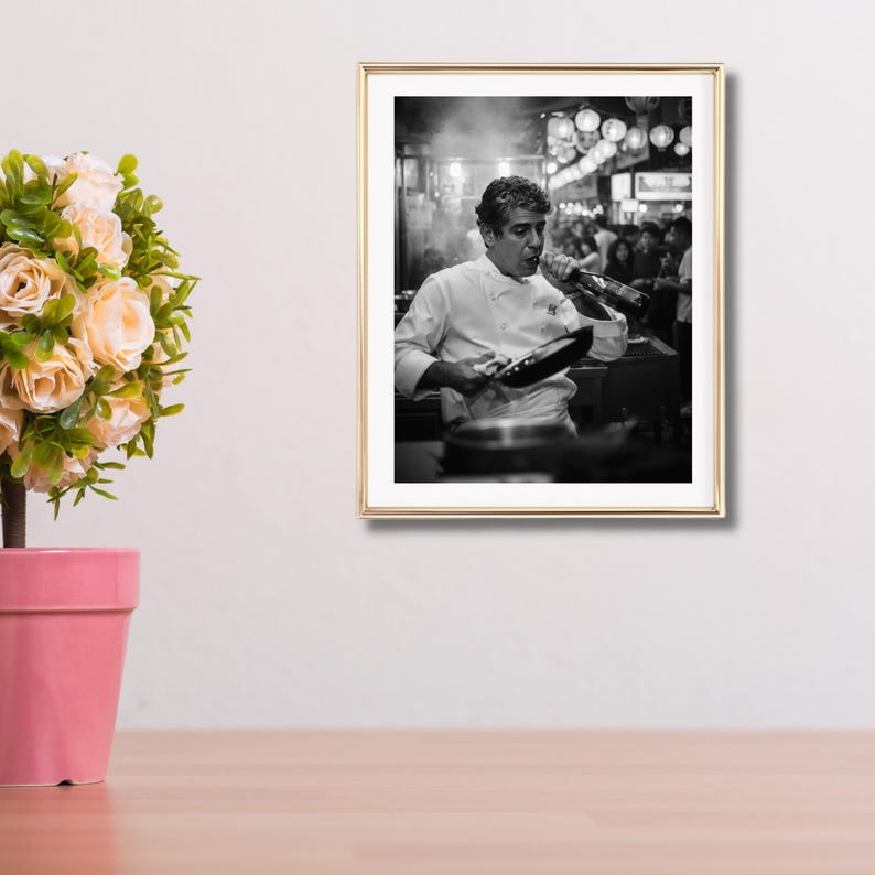 Peut inclure: Photographie encadr&eacute;e en noir et blanc d'un chef en blouse blanche, tenant une po&ecirc;le et une bouteille. La photographie est dans un cadre dor&eacute; et est accroch&eacute;e &agrave; un mur blanc. Un pot de fleurs rose avec des fleurs est au premier plan.
