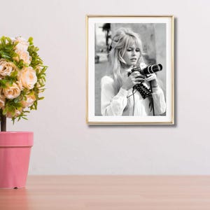 May include: A framed black and white photograph of a woman holding a camera. The image is in a gold frame and shows a woman with long blonde hair and a white blouse. The photograph is a portrait and is displayed on a white wall.