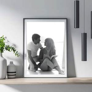 May include: Monochrome framed print depicting a couple in an intimate moment. The woman is seated, reading a book, and the man kneels beside her. The print is on a wooden shelf, with vases and black hanging lights.