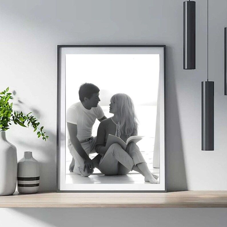 May include: Monochrome framed print depicting a couple in an intimate moment. The woman is seated, reading a book, and the man kneels beside her. The print is on a wooden shelf, with vases and black hanging lights.