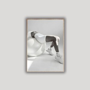 May include: Black and white photograph of a person wearing white athletic shorts, a sports bra, socks, and sneakers, resting on a large white exercise ball. The image is set against a plain white background.