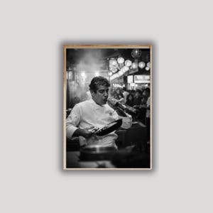 Peut inclure: Photographie en noir et blanc d'un chef en veste blanche, tenant une po&ecirc;le et une bouteille. Le chef souffle dans la bouteille, avec une foule et des lanternes en arri&egrave;re-plan. L'image est encadr&eacute;e dans un cadre en bois.