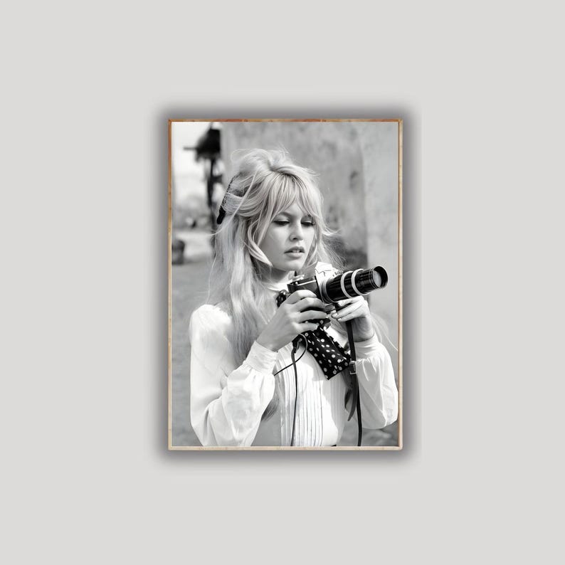 May include: Black and white framed photograph of a woman holding a vintage camera. The woman has long blonde hair and is wearing a white blouse. The camera has a long lens and a black and white polka dot strap.