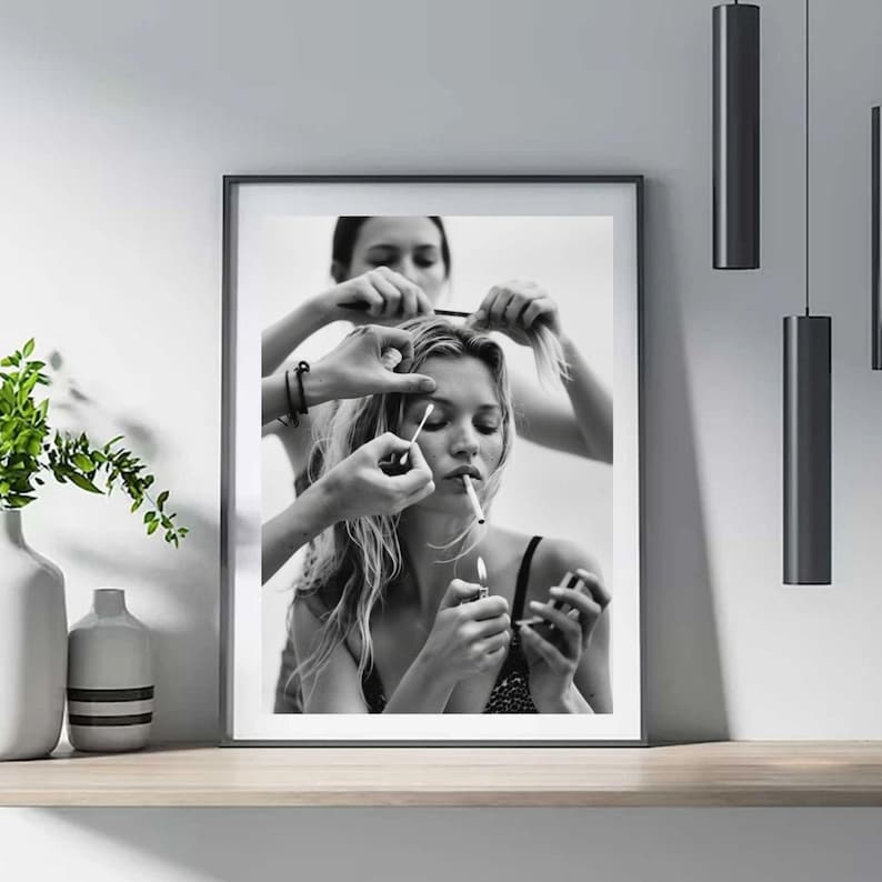 May include: Black and white framed print featuring a woman smoking while having her hair and makeup done. The image shows multiple hands working on her hair and applying makeup. The print is displayed on a wooden shelf.