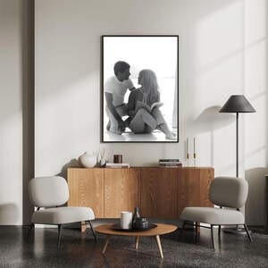 May include: Monochrome framed print of a couple gazing at each other. The print is above a wooden cabinet, a round coffee table, and two grey chairs. A black floor lamp stands to the right.