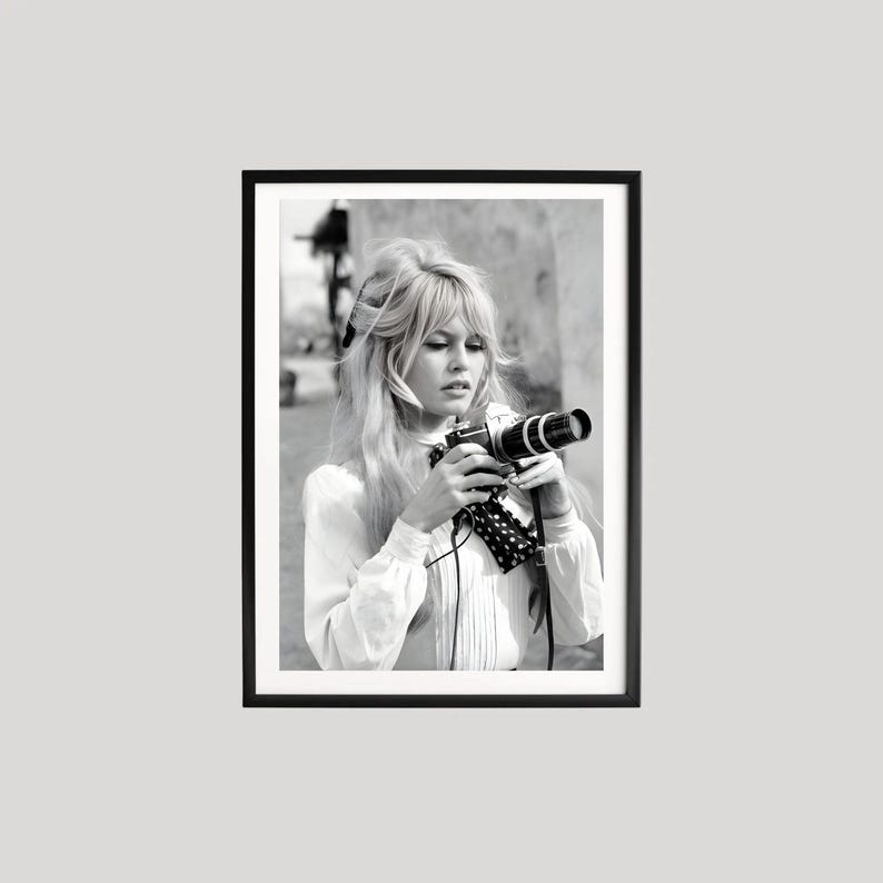 May include: A monochrome framed print featuring a woman holding a vintage camera. The woman has long blonde hair and wears a white blouse with a polka dot scarf. The print is presented in a black frame.