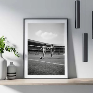 K&ouml;nnte beinhalten: Schwarz-Wei&szlig;-Druck in einem Rahmen, der zwei Frauen in Badeanz&uuml;gen zeigt, die auf einem Fu&szlig;ballfeld rennen, sich an den H&auml;nden halten und einen Fu&szlig;ball haben. Im Hintergrund ist ein Stadion mit Zuschauern zu sehen.