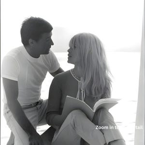 May include: A monochrome photograph of a man and a woman. The woman is reading a book, wearing a light-coloured top and trousers. The man is in a white t-shirt and light-coloured trousers. The image includes the text "Zoom in to detail".