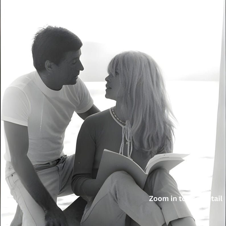 May include: A monochrome photograph of a man and a woman. The woman is reading a book, wearing a light-coloured top and trousers. The man is in a white t-shirt and light-coloured trousers. The image includes the text "Zoom in to detail".